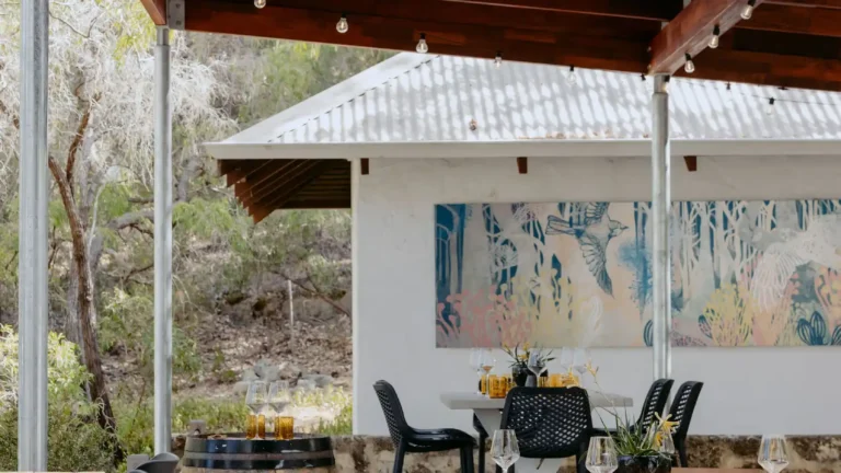 Outdoor dining area at Valle du Venom in Yallingup with round timber table, wine glasses, amber glassware, wine barrel side tables, and large botanical artwork on the back wall