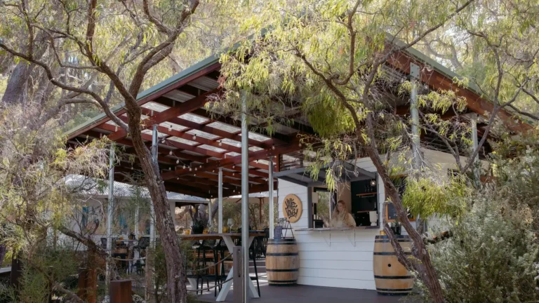 Valle du Venom winery exterior viewed through native Australian trees in Yallingup, with a staff member visible at the cellar door counter and wine barrels on the deck